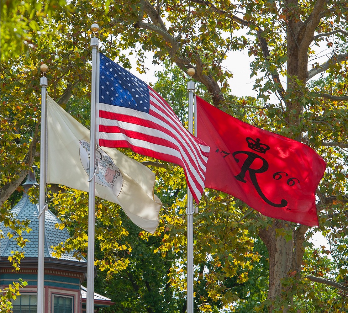 Three flags on poles outdoors—the New Jersey state flag, the United States flag, and a red Rutgers University flag—waving in front of leafy trees on a sunny day.