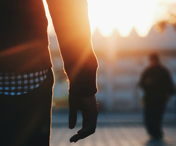 Person standing outdoors at sunset with another person blurred in the background.