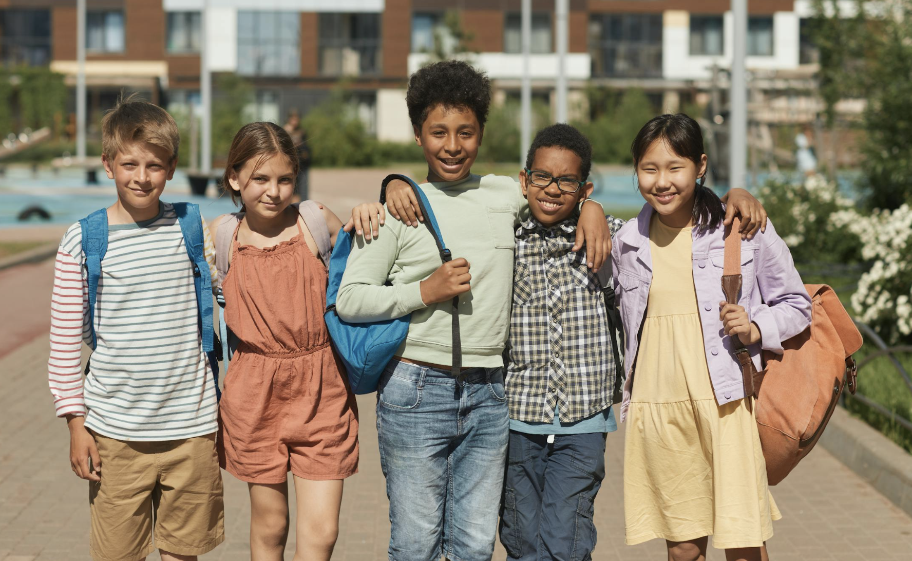 A group of five elementary school students walk together outdoors with backpacks, smiling and supporting each other.