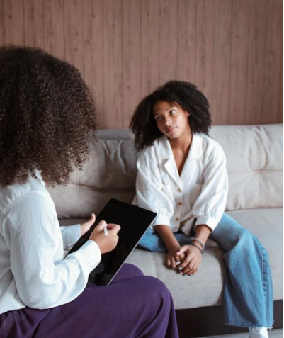 Clinician sitting with a child on a couch during a therapy session, holding a tablet and listening attentively.