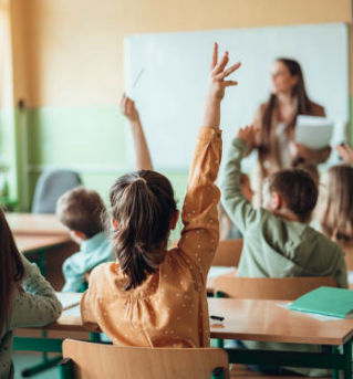 Students in a classroom raising their hands while a teacher leads a lesson at the front of the room.