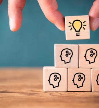 Hand placing a block with a lightbulb icon on top of stacked blocks with head and gear symbols, representing learning and development.