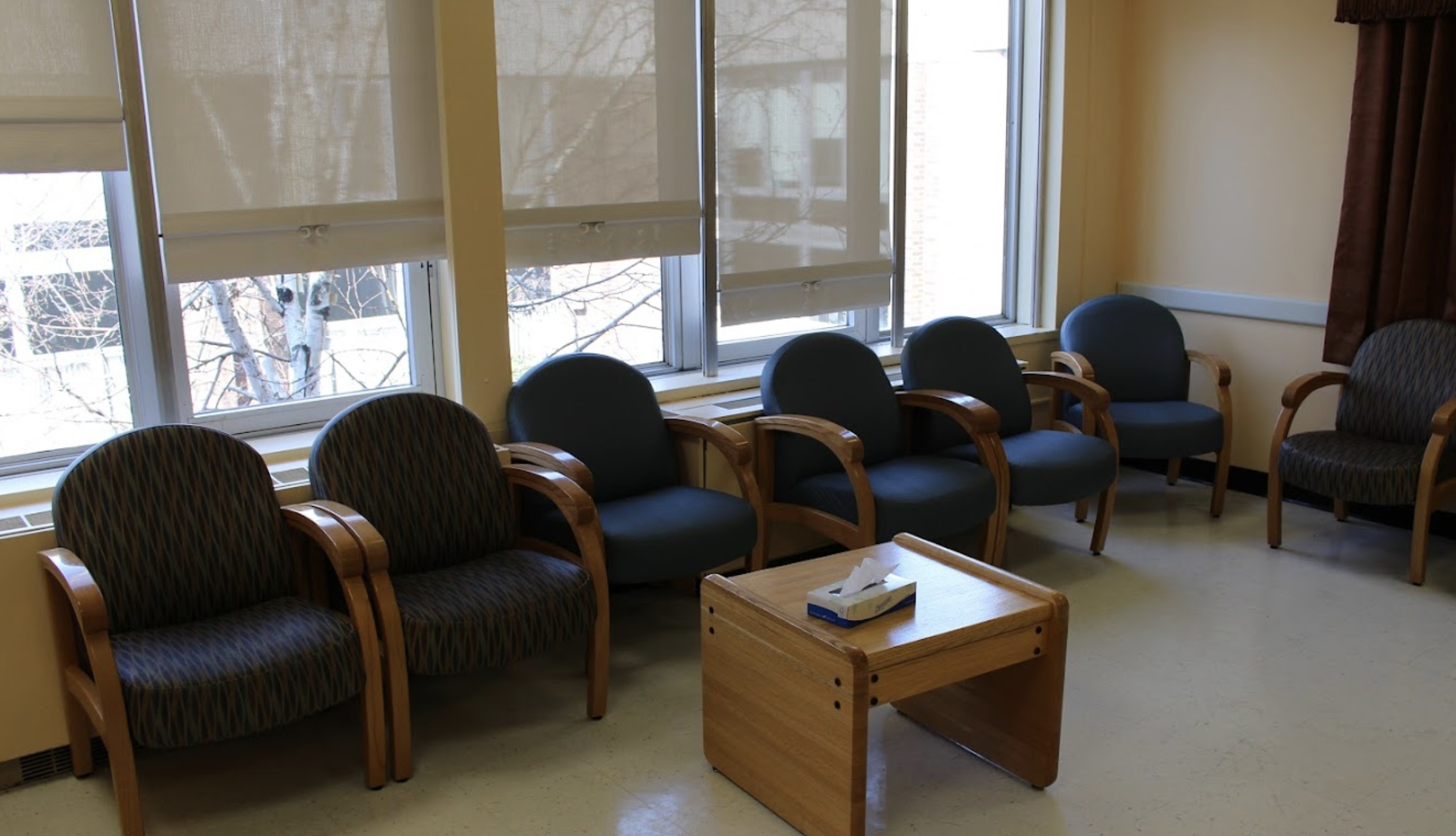 Waiting area within an inpatient behavioral health unit featuring upholstered chairs arranged along large windows with partially lowered shades and a central wooden table with tissues.