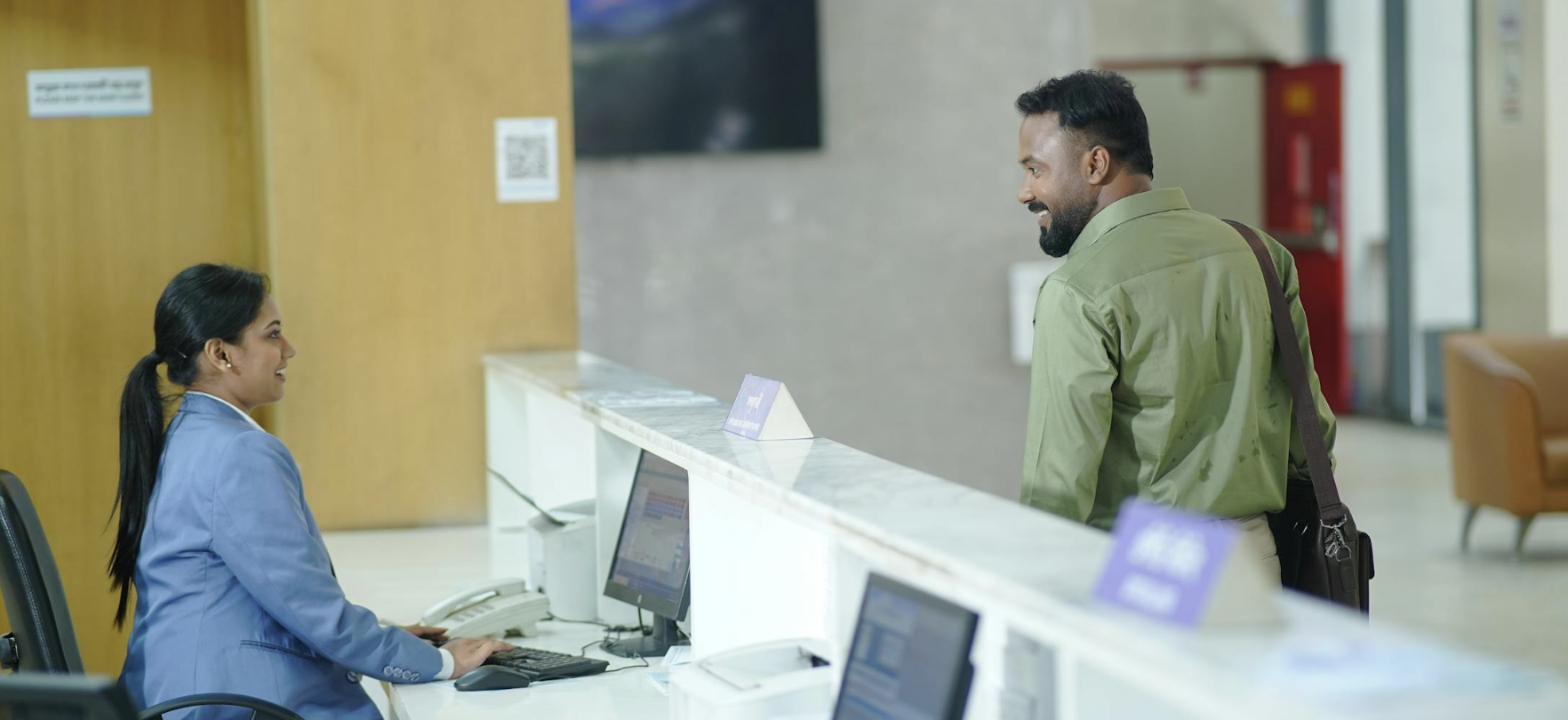 Reception staff member assisting a visitor at a healthcare registration desk in a welcoming outpatient setting.