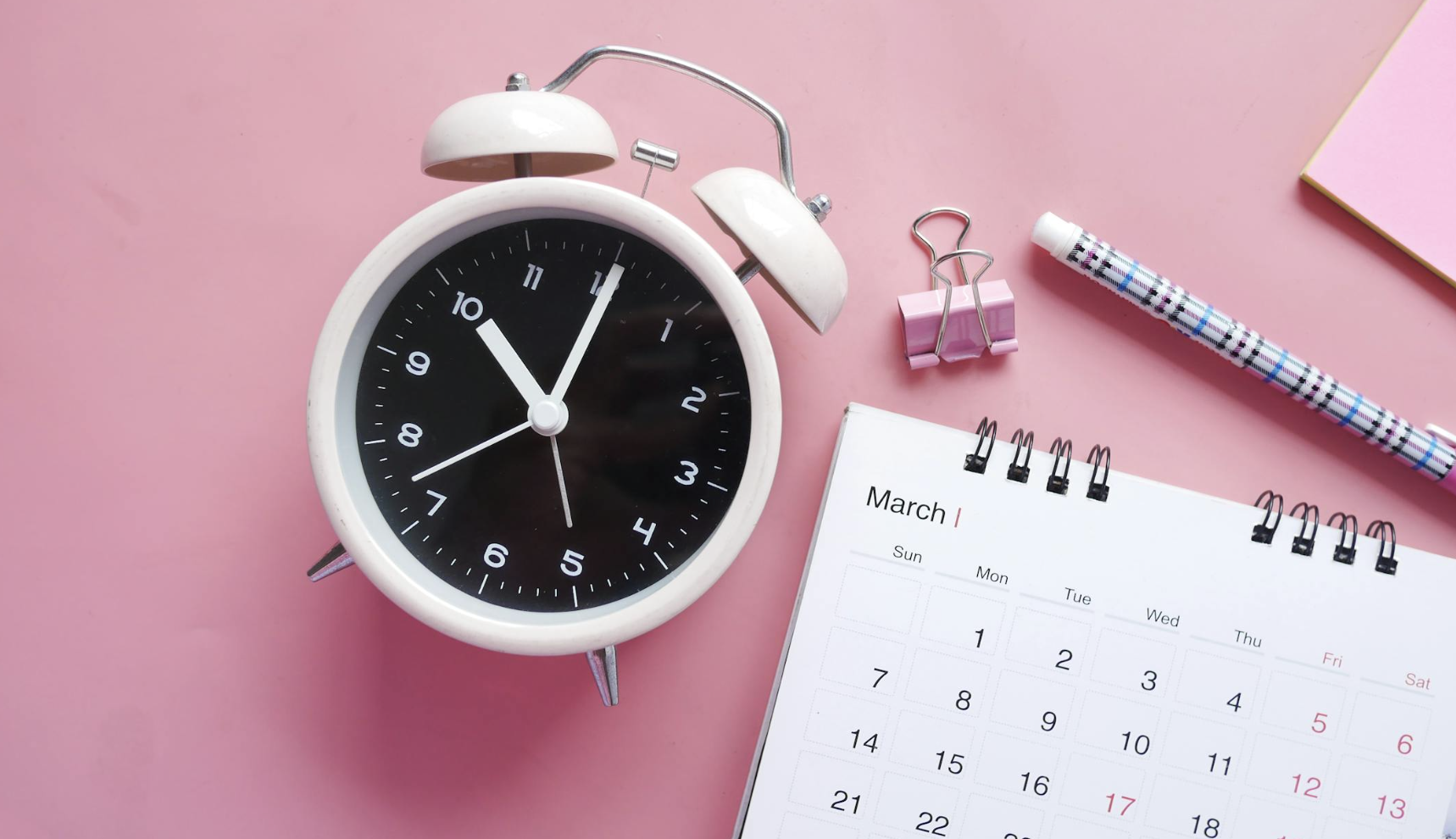 Desk calendar, alarm clock, and office supplies arranged on a pink background, representing scheduling, appointments, and time management.