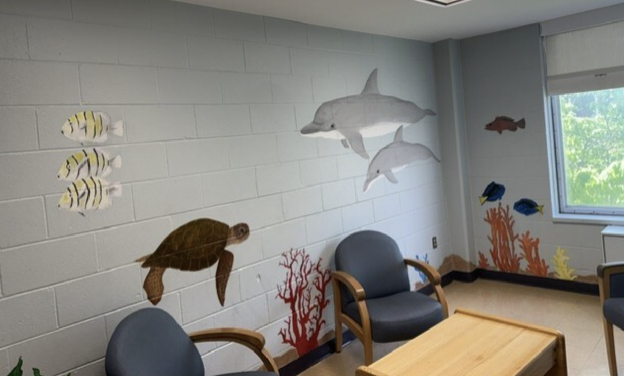 Lounge in an inpatient behavioral health unit featuring ocean-themed wall art with dolphins, fish, a sea turtle, coral, comfortable chairs, natural light, and a small coffee table.