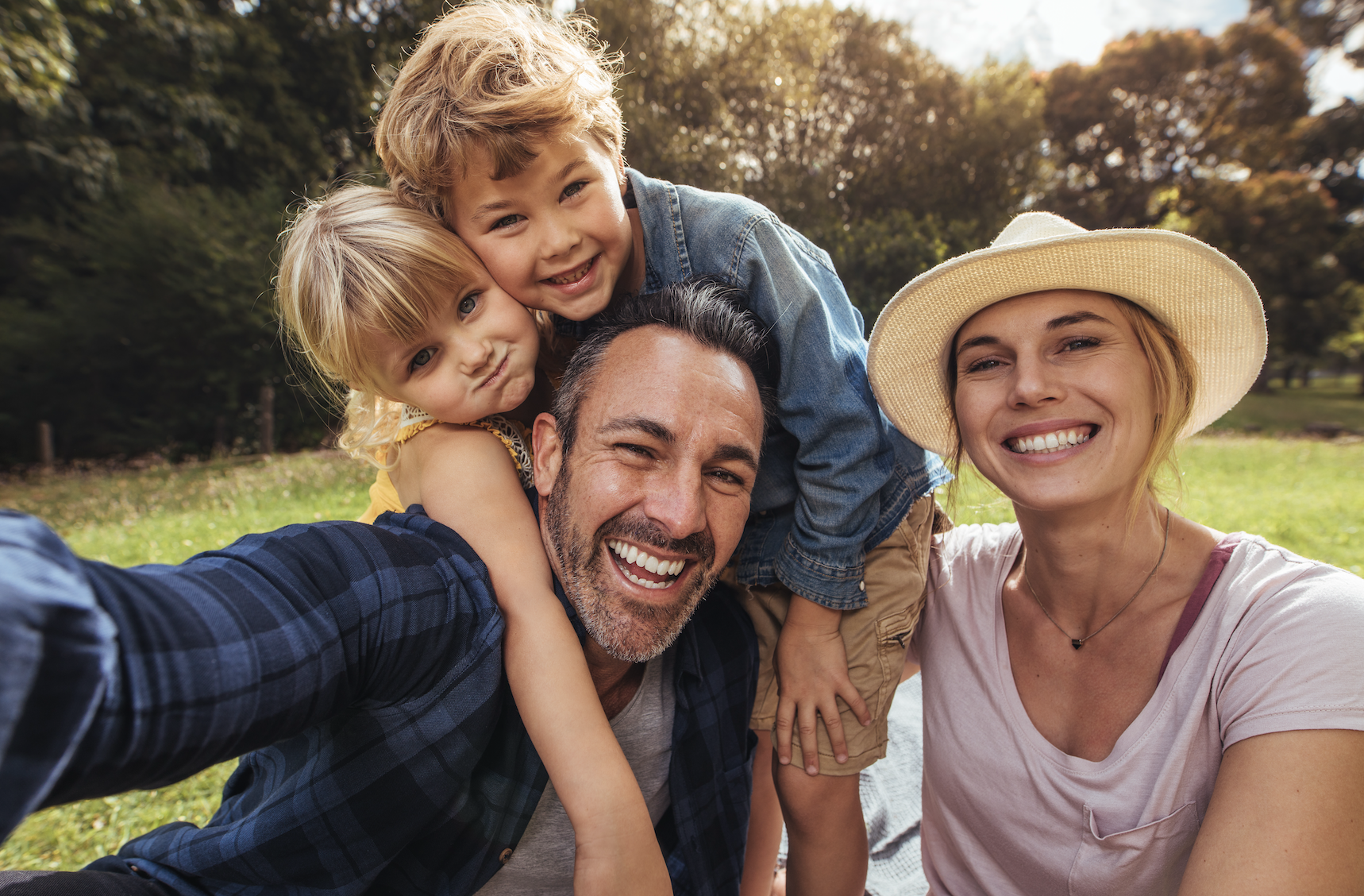 A smiling family of four takes a selfie while enjoying a sunny day together in a park, with two young children hugging their parents.