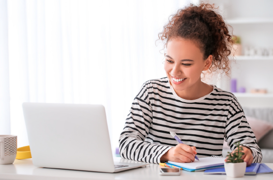 A smiling teenager sits at a desk, writing in a notebook while using a laptop for studying or online learning.