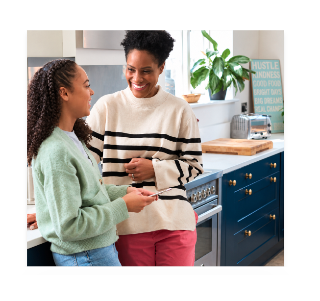 A parent and teenager smile at each other while standing together in a bright kitchen, with the teen holding a smartphone.