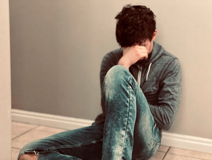 Young person sits on the floor with their head lowered and face covered by one hand, appearing distressed.