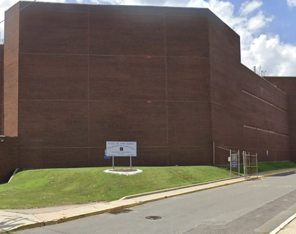 Exterior of a large brick New Jersey correctional facility with a Department of Corrections sign, lawn, and secured entrance.