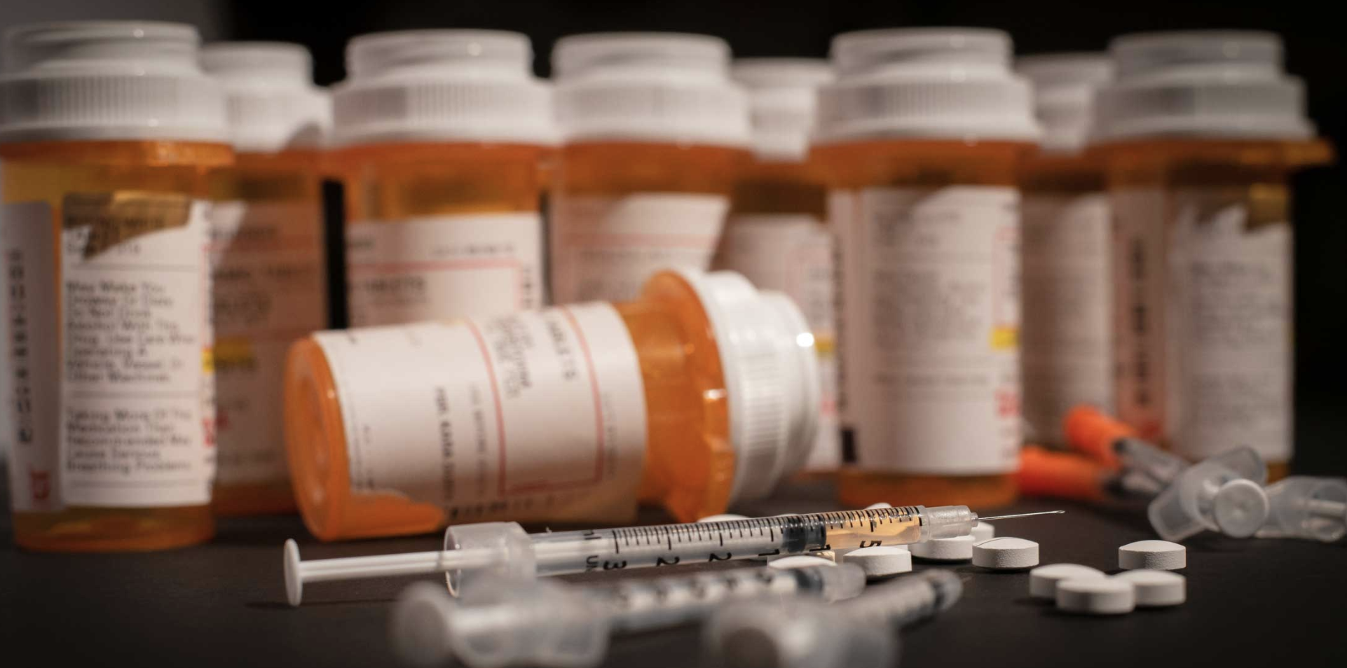 Prescription medication bottles, tablets, and syringes arranged on a dark surface.
