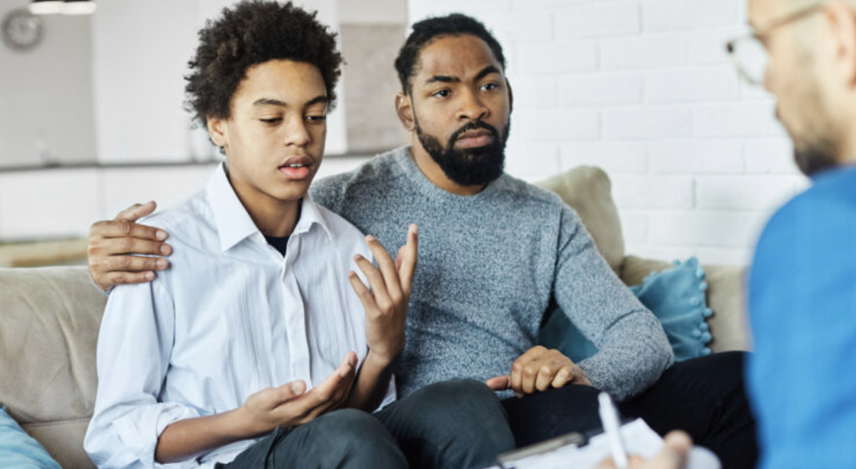 A teenager speaks with a behavioral health professional while an adult caregiver sits beside him with a supportive arm around his shoulder.