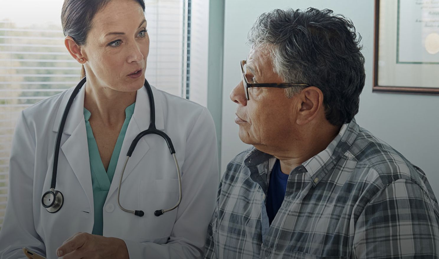 A physician wearing a white coat and stethoscope speaks with an older adult patient during an office visit.