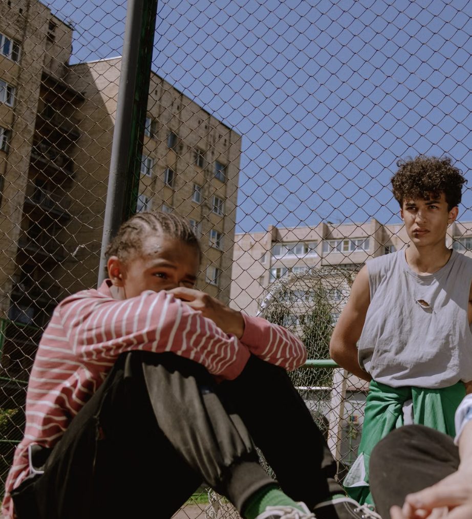 Two young people gathered outdoors near a fenced recreation area, with one seated and another standing nearby.