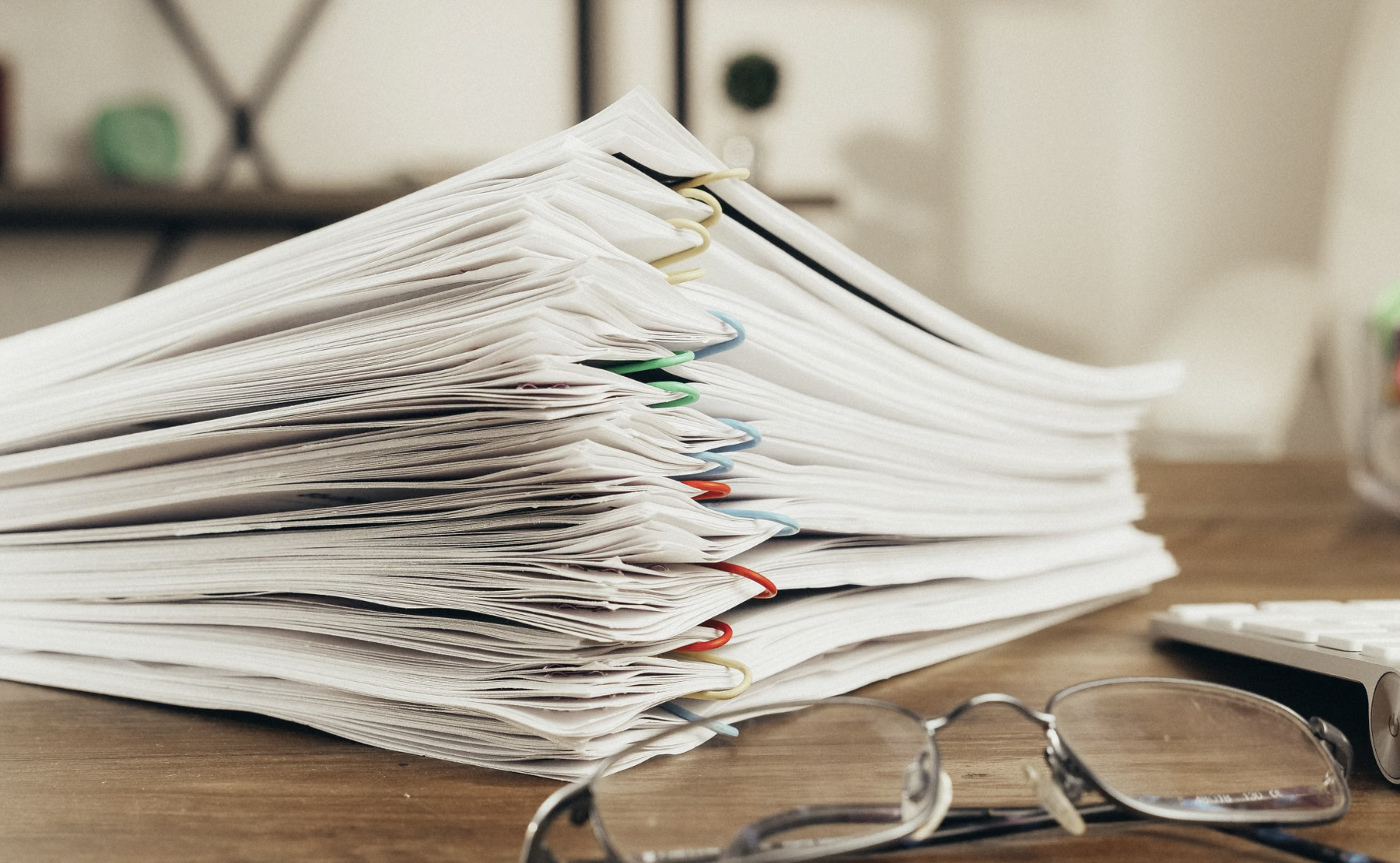 Stack of paperwork and documents secured with colorful paper clips on an office desk, with eyeglasses and a keyboard nearby.
