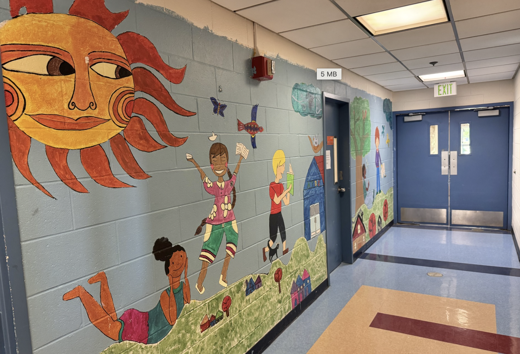 School hallway featuring a colorful wall mural with a large sun, children reading and playing, trees, and neighborhood scenes.