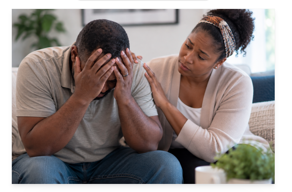 Woman comforts a distressed man sitting with his head in his hands at home.